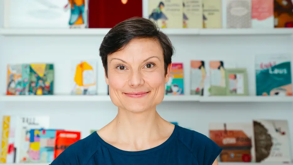 Eine l&auml;chelnde Frau mit kurzen, glatten Haaren vor einem B&uuml;cherregal.