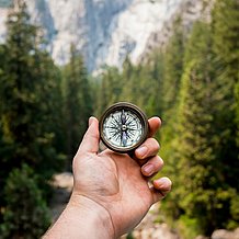 Auf dem Bild ist eine ausgestreckte Hand vor einer Waldlandschaft zu sehen, die einen Kompass hält