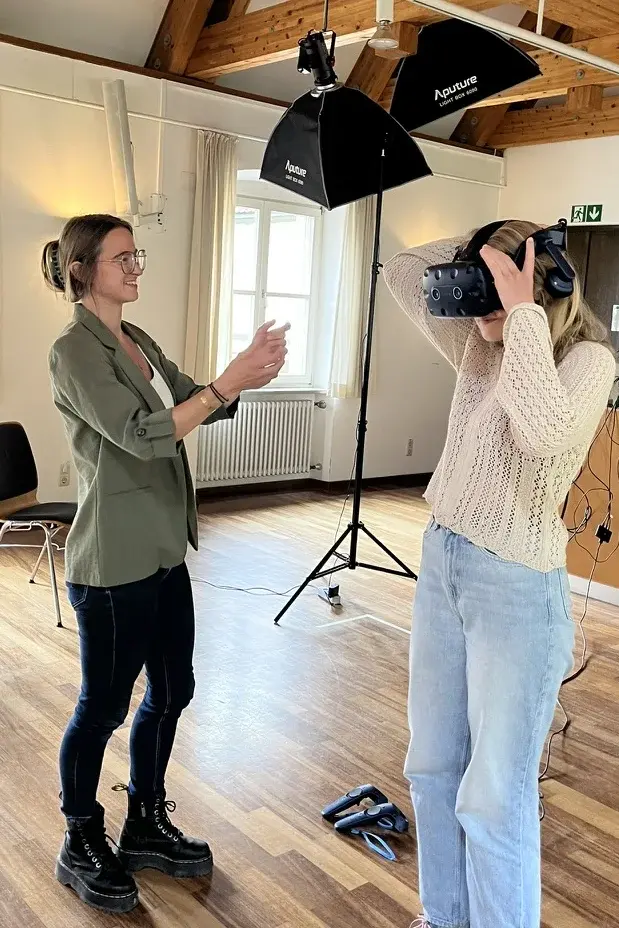 Eine Studierende tr&auml;gt eine VR-Brille und interagiert in einem Seminarraum mit der virtuellen Lernumgebung. Eine weitere Person begleitet die Situation und gibt Hinweise zur Nutzung der Technik.