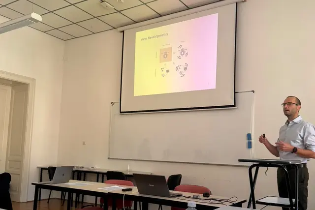 Ein Referent hält in einem Seminarraum einen Vortrag; auf der Leinwand eine Folie mit dem Titel „new developments“.