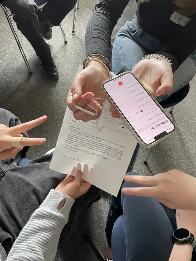 Die Demokratiedetektive nehmen gemeinsam mit einer Studentin das Intro f&uuml;r eine Podcastfolge auf. Zu sehen sind H&auml;nde, ein Smartphone mit laufender Audioaufnahme und ein Arbeitsblatt zum Thema Demokratie in Bayern.