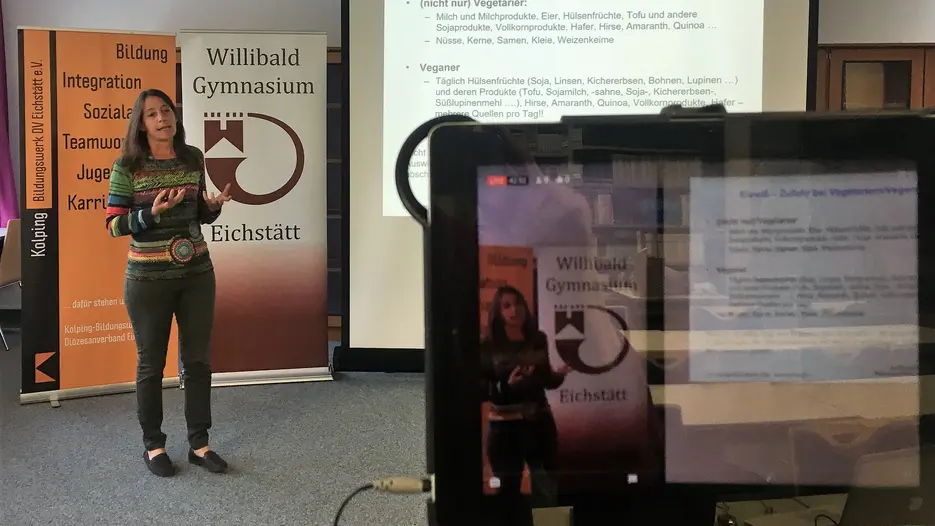 Dr. Daniela Muhr-Becker steht vor Rollups des Kolping-Bildungswerks und des Willibald-Gymnasiums, referiert und zeigt eine Folie, auf der es um die Eiweiß-Zufuhr bei Vegetariern und Veganern geht. Im Vordergrund des Bildes ist ein Tablet-Computer, auf dessen Display man das aktuell live übertragene Bild sehen kann.