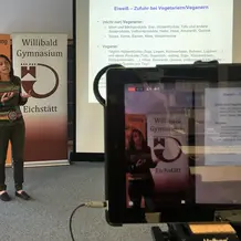 Dr. Daniela Muhr-Becker steht vor Rollups des Kolping-Bildungswerks und des Willibald-Gymnasiums, referiert und zeigt eine Folie, auf der es um die Eiweiß-Zufuhr bei Vegetariern und Veganern geht. Im Vordergrund des Bildes ist ein Tablet-Computer, auf dessen Display man das aktuell live übertragene Bild sehen kann.