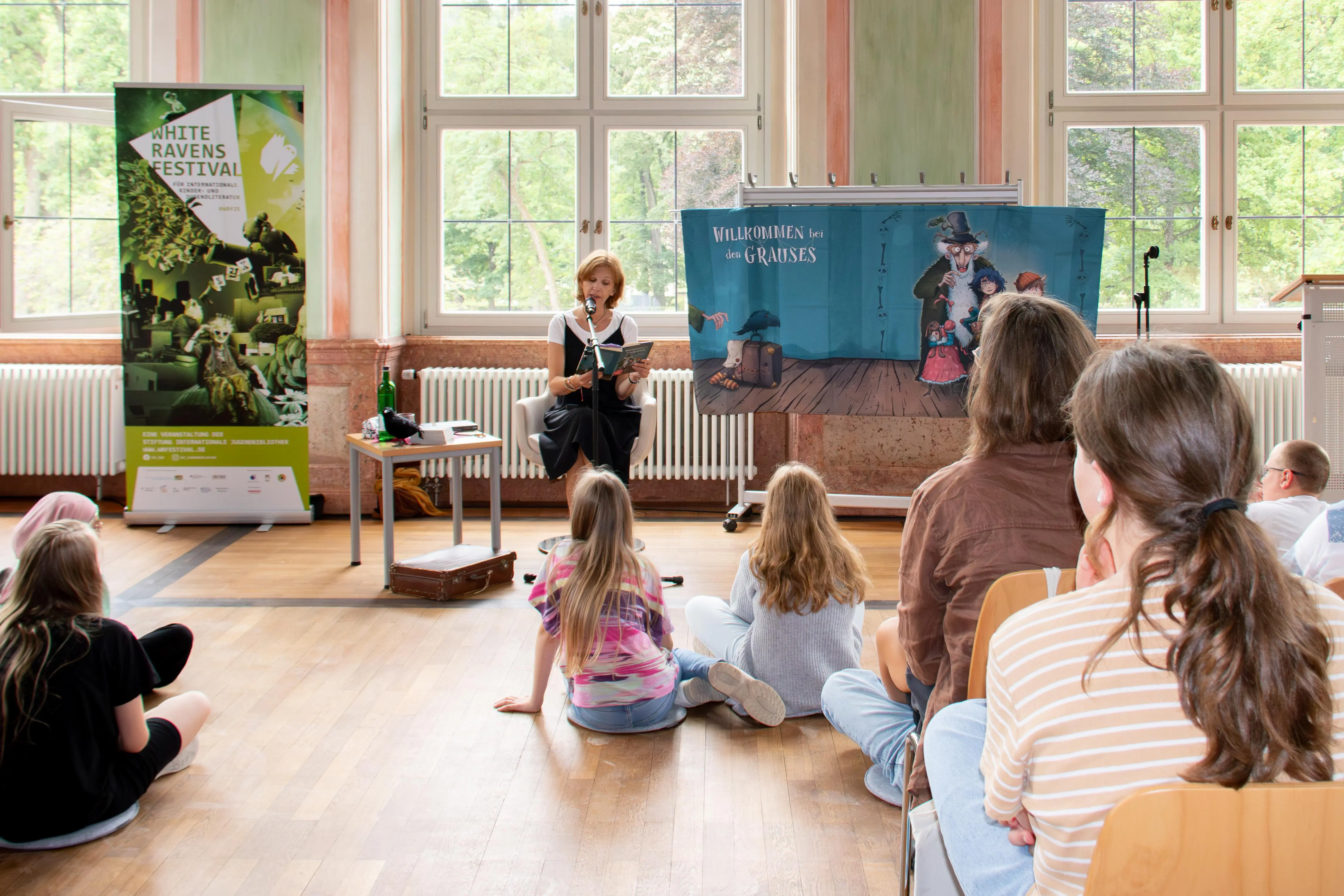 Eine Frau sitzt in einem Sessel und liest aus einem Buch. Vor ihr sitzen Kinder auf Sitzkissen auf dem Boden.
