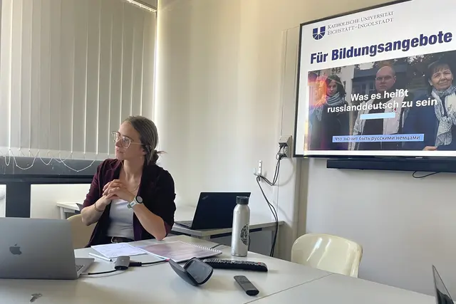 Lisa Schmidt sitzt w&auml;hrend einer Pr&auml;sentation an einem Tisch in einem Seminarraum. Vor ihr liegen ein Laptop und Unterlagen. Im Hintergrund ist eine Pr&auml;sentationsfolie der Katholischen Universit&auml;t Eichst&auml;tt-Ingolstadt zum Thema &bdquo;Was es hei&szlig;t, russlanddeutsch zu sein&ldquo; zu sehen.
