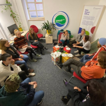 Junge Menschen sitzen im Kreis auf Stühlen in einem Raum, in dem ein Schild mit "Green Office" steht 