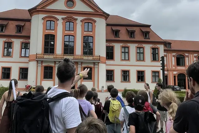 Eine Gruppe von F&uuml;nftkl&auml;ssler*innen steht bei einer Campusf&uuml;hrung vor der Sommerresidenz der KU Eichst&auml;tt-Ingolstadt. Im Vordergrund zeigt Prof. Dr. Rico Behrens auf das historische Geb&auml;ude.