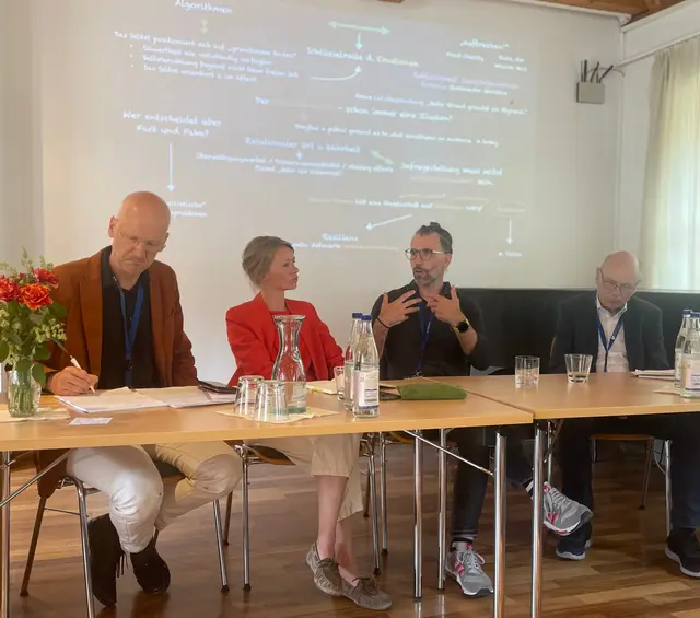 Vier Personen sitzen beim Abschlusspodium der 3. ZRKG-Tagung an einem Tisch in einem Seminarraum. Im Hintergrund ist eine projizierte Tagungszusammenfassung zu sehen. Rico Behrens spricht, w&auml;hrend Martin Kirschner, Imke von Maur und Hans-Joachim H&ouml;hn zuh&ouml;ren.