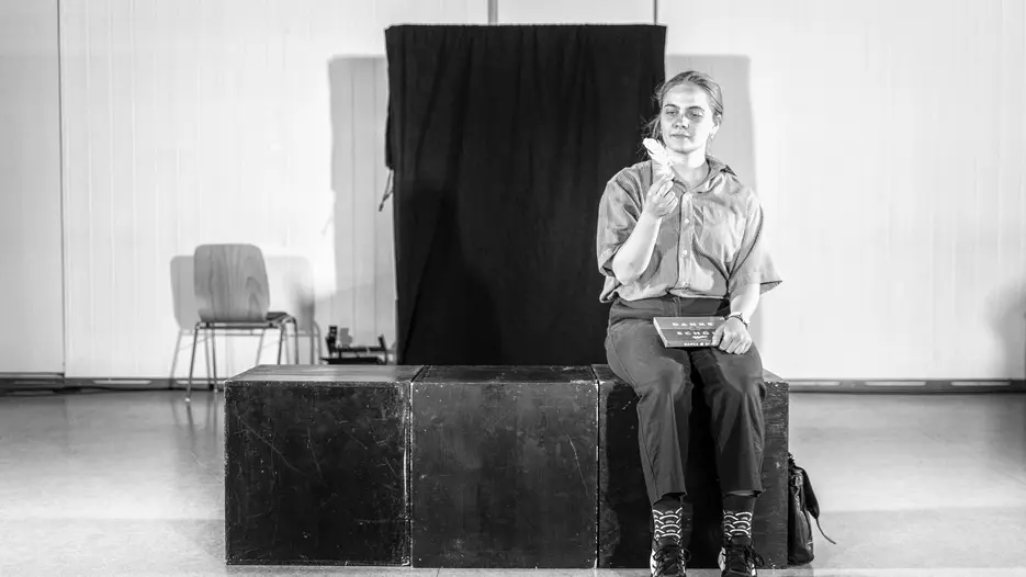 Forrest Gump - man weiß nie, was man bekommt. Eine Spielszene: Eine junge Frau mit zurückgebundenen Haaren sitzt auf einer Kiste, im Schoß eine Schachtel Pralinen, in der rechten Hand eine weiße Vogelfeder, die sie versonnen betrachtet.