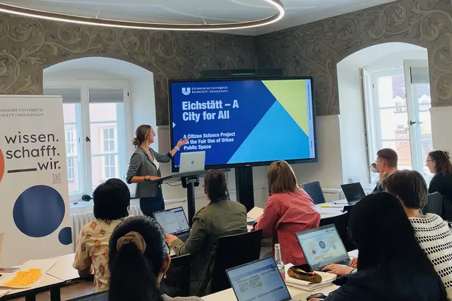 Lisa Schmidt stellt das Citizen-Science-Projekt &bdquo;Eichst&auml;tt &ndash; A City for All&ldquo; vor. W&auml;hrend der Pr&auml;sentation erl&auml;utert sie am Bildschirm das Projekt zur fairen Nutzung des &ouml;ffentlichen Raums; im Vordergrund sind Teilnehmende des Workshops zu sehen.