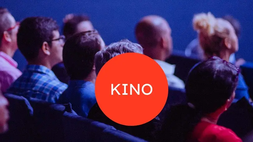 Im Hintergrund sieht man Kinopublikum, vorne ist ein roter Kreis zu sehen, in dessen Mitte in weißer Schrift "KINO" steht.