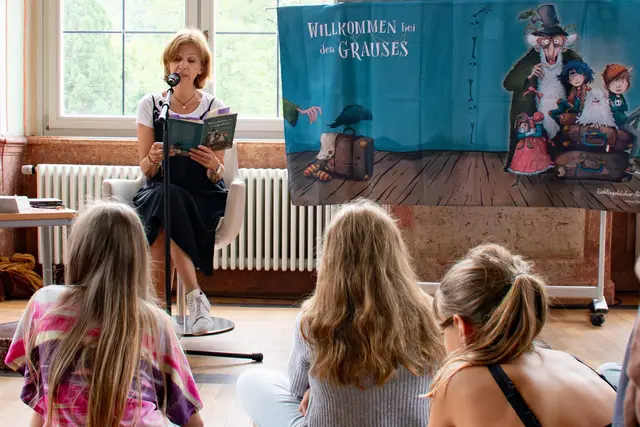Eine Frau sitzt in einem Sessel und liest aus einem Buch. Neben ihr hängt ein buntes Banner mit Kinderbuchfiguren. Vor ihr sitzen Kinder auf dem Fußboden und hören zu.