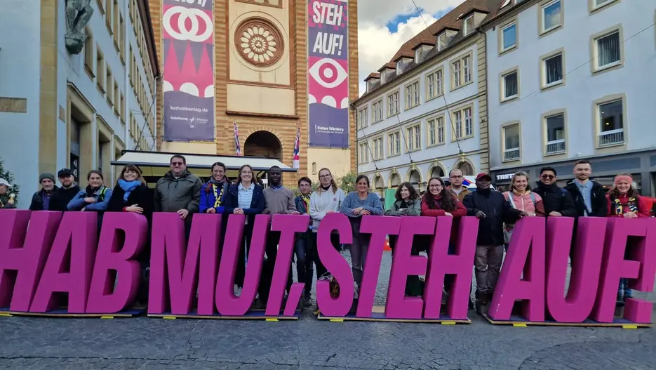 Die 16 teilnehmenden Studierenden mit Prof. Dr. Marievonne Sch&ouml;ttner, Dr. Tanja Grabovac und Prof. Dr. Katharina Karl (von links nach rechts) mit Katholikentagsmotto und Kiliansdom in W&uuml;rzburg